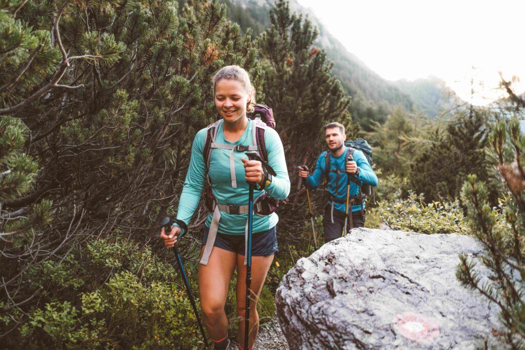 Smiling woman hiker and her partner walking behind her on a beautiful trail up in the Alps early in the morning on a summer day